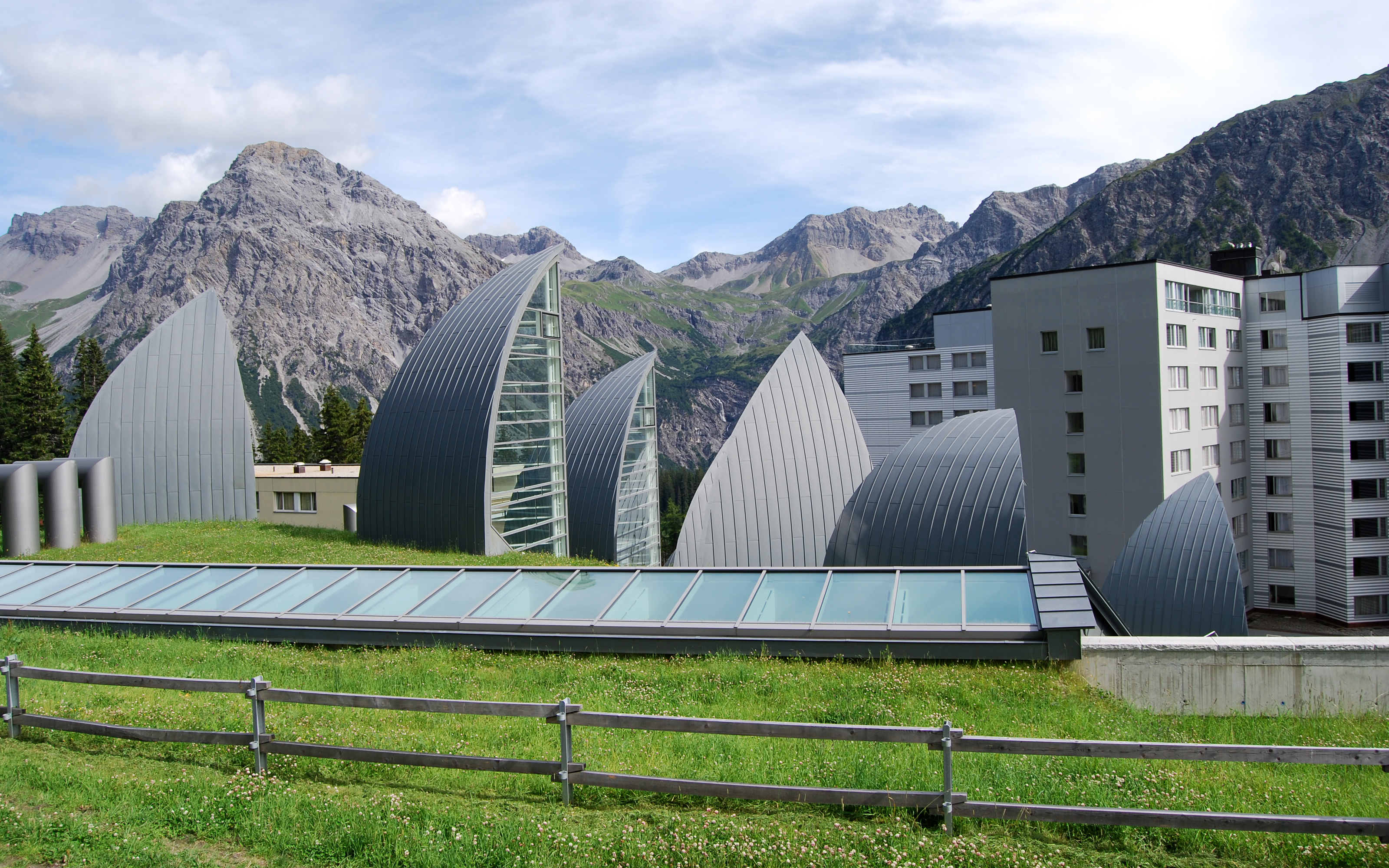 The design simulates its mountainous surroundings. Sail-shaped skylights on a green roof in front of mountains